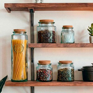 May include: A collection of glass jars with cork lids, filled with various dry goods, displayed on wooden shelves. The jars contain pasta, coffee beans, salt, peppercorns, and herbs. The jars have an embossed design and are arranged on a wooden shelf.