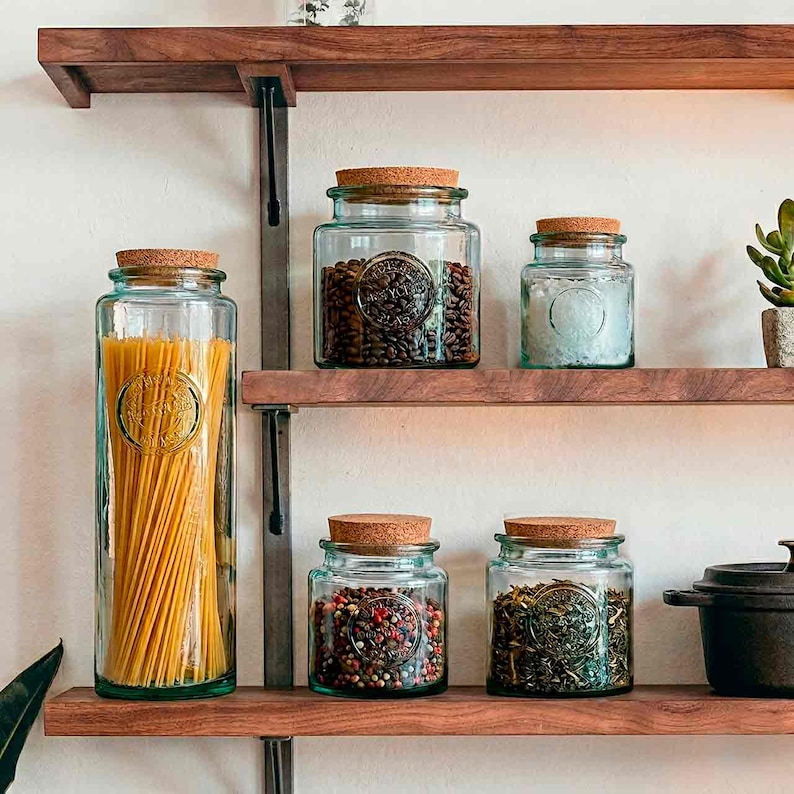 May include: A collection of glass jars with cork lids, filled with various dry goods, displayed on wooden shelves. The jars contain pasta, coffee beans, salt, peppercorns, and herbs. The jars have an embossed design and are arranged on a wooden shelf.