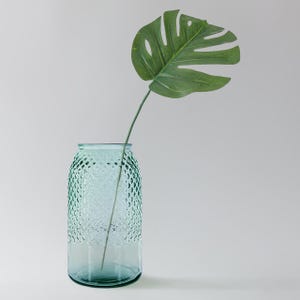 May include: A teal blue glass vase with a textured surface, holding a single green leaf.