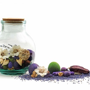 May include: A glass jar with a cork lid filled with potpourri. The potpourri includes dried flowers, lavender, and other natural elements. The jar is sitting on a white surface with a scattering of lavender buds.