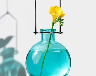 May include: A round, turquoise glass vase with a black metal hanger. A yellow flower with green stem is placed inside the vase. The vase is filled with water. The background is white.