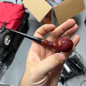 Peut inclure: Gros plan d'une main tenant une pipe en bois brun foncé et noir avec une coupe texturée. La pipe mesure environ 15 cm de long.