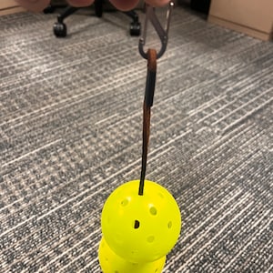 Puede incluir: Un llavero de pickleball amarillo con dos pelotas perforadas apiladas. Las pelotas están unidas a una correa de cuero marrón y un mosquetón plateado.