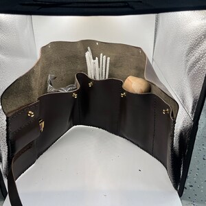 May include: A brown leather tool roll with multiple pockets and compartments. The roll is open and contains white pipe cleaners, a small plastic bag, and a wooden dowel.