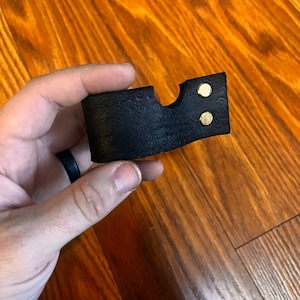 May include: A black leather case with a decorative embossed pattern and two gold metal accents. The case has a cut-out for a small object.