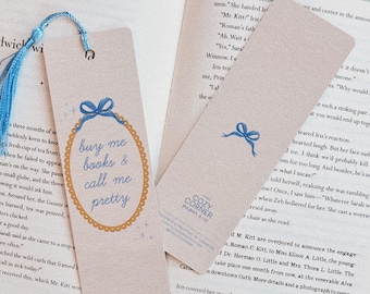 May include: A blue and gold bookmark with the text "buy me books & call me pretty" in a decorative oval frame. The bookmark has a blue ribbon with a bow at the top.