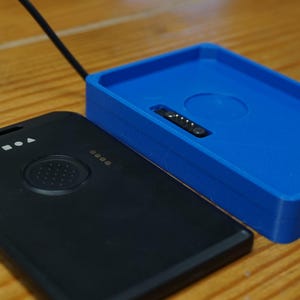 May include: A black rectangular device with a circular button and indicator lights, next to a bright blue rectangular tray. A black cord extends from the device. The items are on a wooden surface.