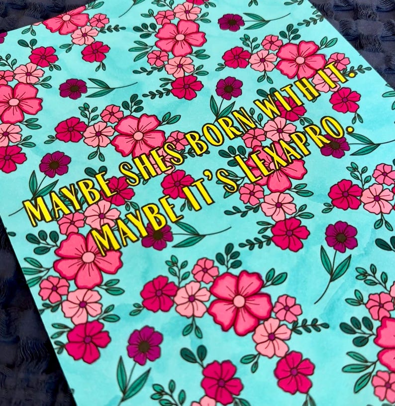 May include: A light blue floral print with pink and magenta flowers and green leaves. The text "MAYBE SHE'S BORN WITH IT. MAYBE IT'S LEXAPRO." is in yellow.