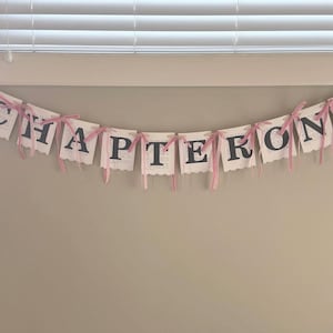 May include: A decorative banner featuring ten individual white paper cards, each printed with a single black letter to spell out the words CHAPTER ONE. The cards are connected by thin, light pink ribbons tied in bows between each letter.