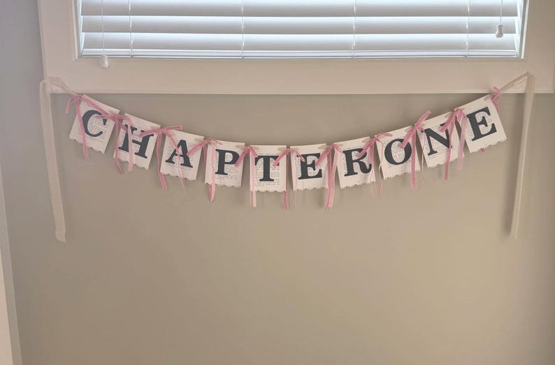 May include: A decorative banner featuring ten individual white paper cards, each printed with a single black letter to spell out the words CHAPTER ONE. The cards are connected by thin, light pink ribbons tied in bows between each letter.