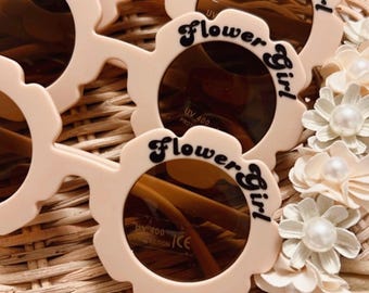 May include: Pink flower-shaped sunglasses with brown lenses and the text "Flower Girl" on the frames.