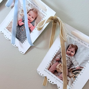 May include: A milestone photo banner featuring two white paper flags with scalloped edges, each displaying a baby photograph and printed text. The left flag is tied with a light blue velvet ribbon, and the right flag is tied with a tan velvet ribbon.