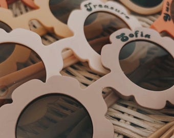May include: A close-up of a collection of colourful flower-shaped sunglasses for children. Each pair has a different name written on the frame, including "Sofia", "Treasure", "Anaish", and "Zander".