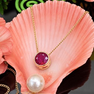 May include: A gold chain necklace featuring a round, bezel-set, faceted deep pink gemstone pendant. The pendant is centered on a light pink seashell, which also holds a single, smooth, round white pearl near the bottom edge of the shell.