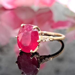 May include: A gold ring with a large, oval-shaped ruby gemstone set in a prong setting. The ruby is a deep red color and has a high polish. The ring also features smaller diamonds set in the band.