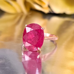 May include: A rose gold ring featuring a large, oval-cut ruby gemstone. The ruby is a vibrant shade of red, and the ring's band has a delicate, polished finish. The ring is set on a reflective surface.