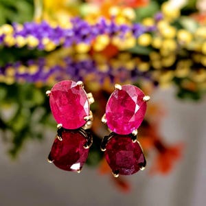 May include: A pair of oval-cut, vibrant pinkish-red gemstone stud earrings set in gold prongs. The faceted stones exhibit a brilliant clarity and deep saturation, reflecting light across their surfaces while resting on a reflective dark base.