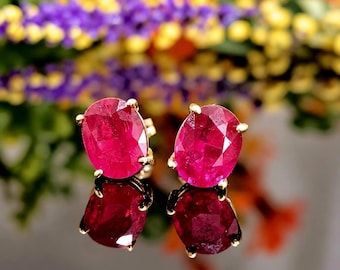May include: A pair of oval-cut, vibrant pinkish-red gemstone stud earrings set in gold prongs. The faceted stones exhibit a brilliant clarity and deep saturation, reflecting light across their surfaces while resting on a reflective dark base.