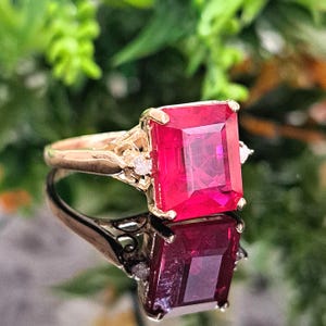 May include: A gold ring featuring a large, rectangular, vibrant red gemstone. The ring has a detailed filigree design on the band and is accented with small, clear gemstones on either side of the main stone. The ring is displayed on a reflective surface.