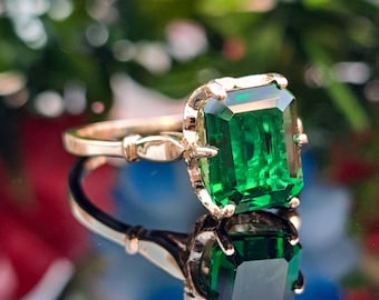 May include: A gold ring with a large, rectangular emerald-cut green gemstone. The ring is set in a gold band with intricate detailing. The gemstone is highly reflective, and the ring is displayed on a reflective surface.