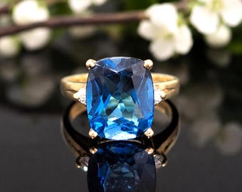 May include: A gold ring featuring a large, cushion-cut, deep blue gemstone as the centerpiece. The ring is accented with a small, clear, triangular-cut stone on each side of the main setting, held in place by gold prongs on a polished gold band.