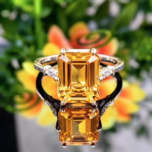 May include: A silver-toned ring featuring a large, rectangular emerald-cut yellow gemstone as the centerpiece. The band is accented on each side by a small, pear-shaped clear stone set in metal, creating a symmetrical design with a polished finish.