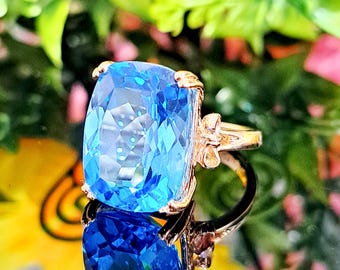 May include: A ring featuring a large, cushion-cut, bright blue gemstone set in a polished rose gold band. The setting includes small, decorative bow-shaped accents on either side of the central stone, which is held in place by four prongs.
