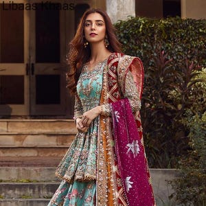 May include: A woman wearing a teal blue and gold embroidered dress with a pink and red dupatta. The dress has a layered skirt and a fitted bodice. The dupatta is draped over the woman's shoulders and is decorated with floral patterns.
