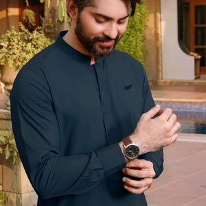 Pode incluir: Uma camisa de manga comprida azul-escura com colarinho mandarim e um pequeno logotipo preto no lado direito. A camisa está abotoada e a pessoa está usando um relógio de couro marrom.