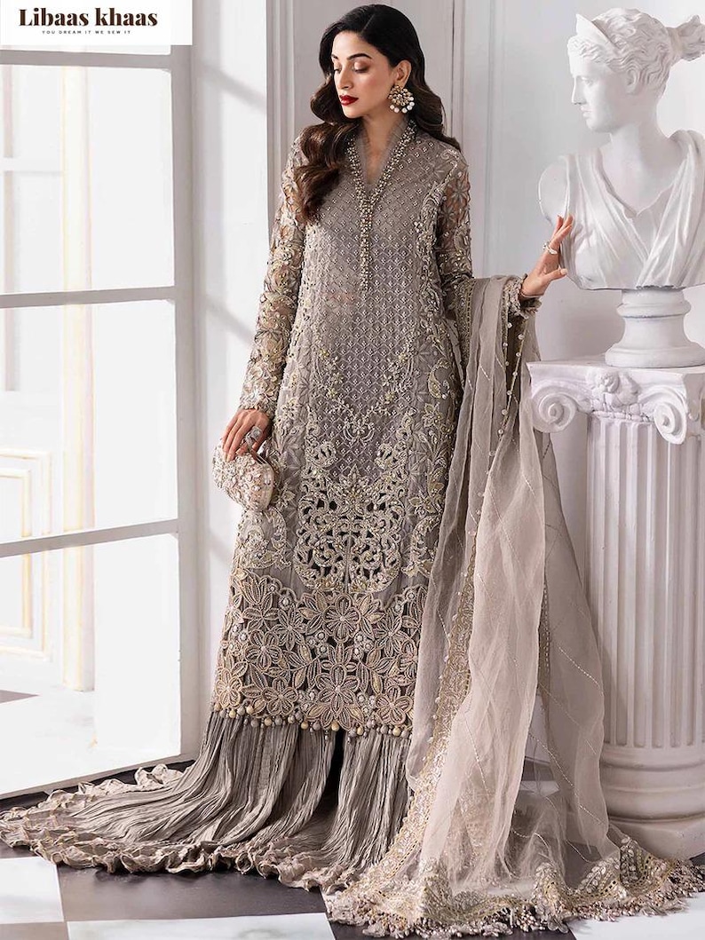 May include: A long, gray embroidered formal dress with intricate floral patterns and bead embellishments. The dress features long sleeves, a flowing skirt, and a matching sheer scarf. A small, embellished clutch is held at the waist.
