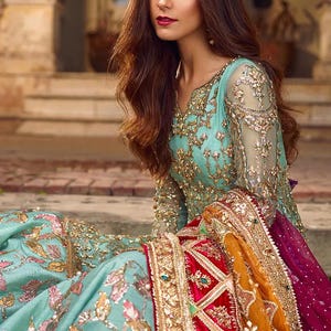 May include: A woman wearing a teal blue and gold embroidered dress with a red and gold dupatta. The dupatta has a velvet border and intricate embroidery.