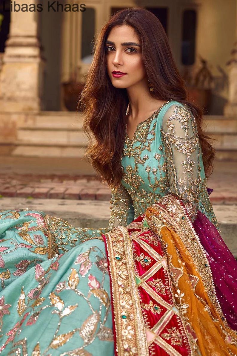 May include: A woman wearing a teal blue and gold embroidered dress with a red and gold dupatta. The dupatta has a velvet border and intricate embroidery.