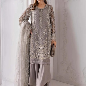 May include: A gray embroidered formal dress with long sleeves and matching pants. The dress features intricate gold detailing and a beaded neckline. A matching scarf and clutch complete the ensemble. The brand name "Libaas Khaas" is visible in the top left corner.