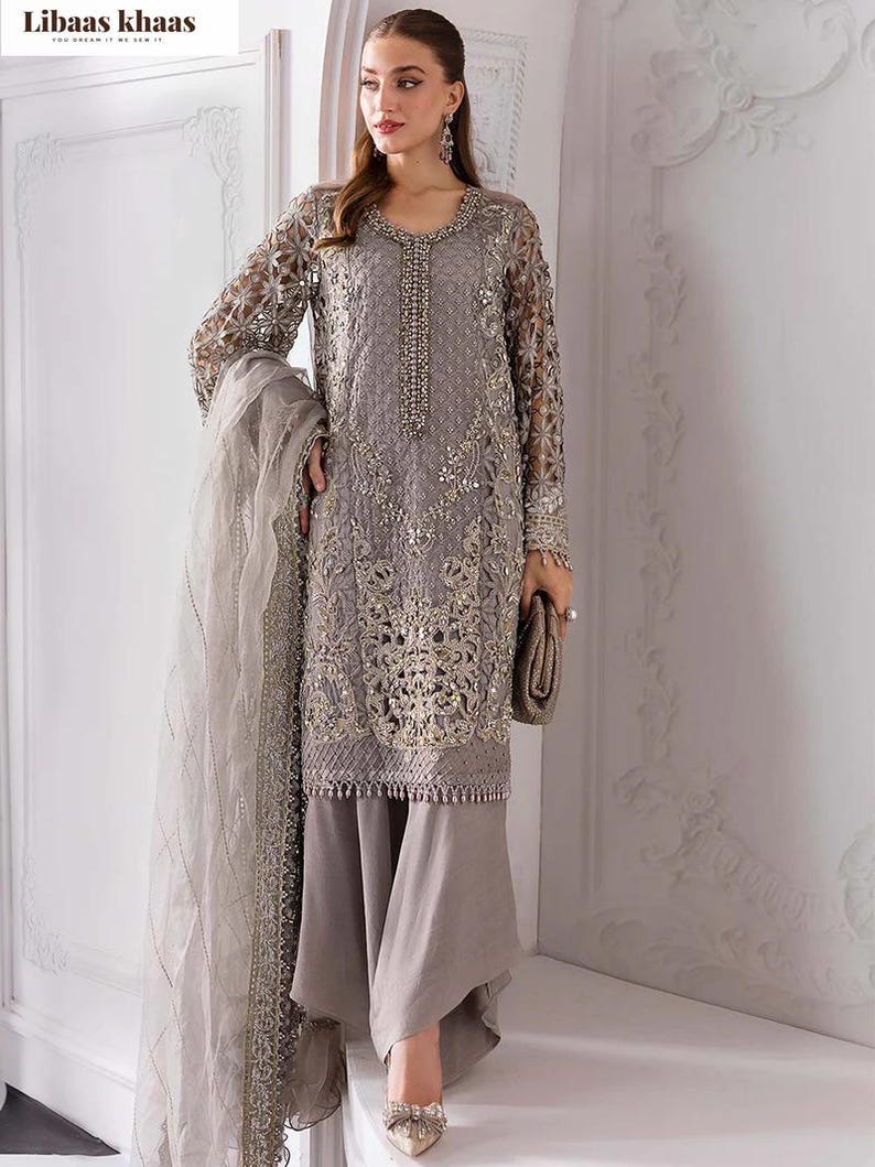 May include: A gray embroidered formal dress with long sleeves and matching pants. The dress features intricate gold detailing and a beaded neckline. A matching scarf and clutch complete the ensemble. The brand name "Libaas Khaas" is visible in the top left corner.