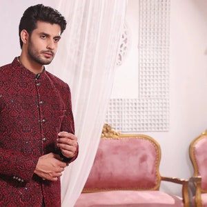 May include: A man wearing a red and black patterned jacket with a mandarin collar. The jacket has a button closure and is made of a textured fabric.