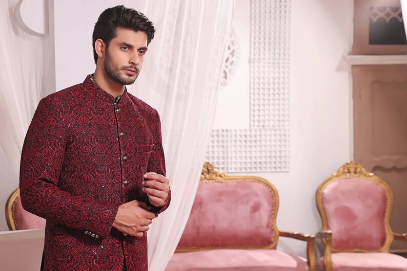 May include: A man wearing a red and black patterned jacket with a mandarin collar. The jacket has a button closure and is made of a textured fabric.