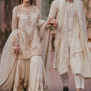 Pu&ograve; includere: Una coppia in abiti formali color crema abbinati. La donna indossa un abito a maniche lunghe con ricami dorati e un copricapo abbinato. L'uomo indossa un cappotto lungo e pantaloni. Entrambi si tengono per mano. L'immagine &egrave; stata scattata all'aperto.