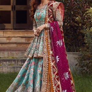May include: A woman wearing a teal blue and gold embroidered dress with a pink and red dupatta. The dress has a layered skirt and a fitted bodice. The dupatta is draped over the woman's shoulders and is decorated with floral patterns.