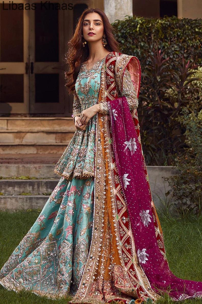 May include: A woman wearing a teal blue and gold embroidered dress with a pink and red dupatta. The dress has a layered skirt and a fitted bodice. The dupatta is draped over the woman's shoulders and is decorated with floral patterns.