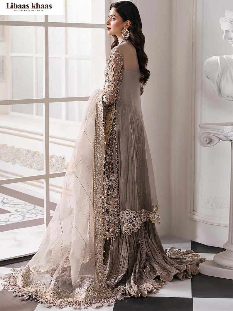 May include: A long, flowing, gray dress with intricate embroidery and a sheer overlay. The dress features detailed lace trim and a matching sheer scarf. The sleeves are adorned with delicate patterns. The dress is suitable for formal occasions.