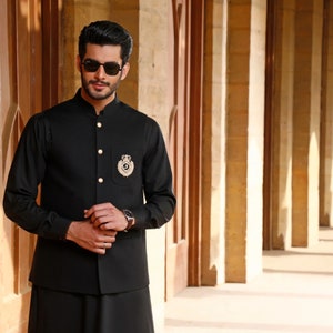 May include: A man wearing a black waistcoat with gold buttons and a gold emblem over a black shirt. He is wearing sunglasses and standing in a hallway with columns.