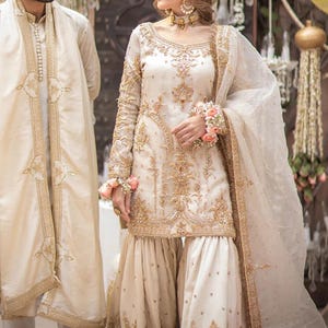 Pu&ograve; includere: Abito da sposa tradizionale color crema con ricami dorati. L'ensemble include una tunica a maniche lunghe, pantaloni a gamba larga e una sciarpa trasparente. Un uomo in un abito abbinato &egrave; accanto alla donna. Il marchio "Libaas Khaas" &egrave; visibile.