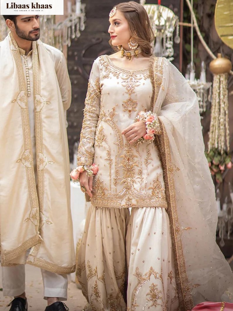 Pu&ograve; includere: Abito da sposa tradizionale color crema con ricami dorati. L'ensemble include una tunica a maniche lunghe, pantaloni a gamba larga e una sciarpa trasparente. Un uomo in un abito abbinato &egrave; accanto alla donna. Il marchio "Libaas Khaas" &egrave; visibile.