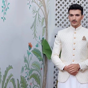 May include: A cream-coloured jacket with a textured pattern and gold buttons. The jacket has a mandarin collar and a pocket square. The person is wearing white trousers. The background features floral wall art.