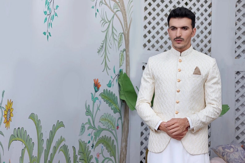 May include: A cream-coloured jacket with a textured pattern and gold buttons. The jacket has a mandarin collar and a pocket square. The person is wearing white trousers. The background features floral wall art.
