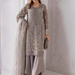 May include: A gray embroidered formal dress with long sleeves and matching pants. The dress features intricate gold detailing and a beaded neckline. A matching scarf and clutch complete the ensemble. The brand name "Libaas Khaas" is visible in the top left corner.