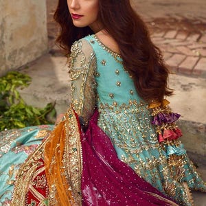 May include: A woman wearing a teal blue and gold embroidered dress with a long, flowing burgundy and orange dupatta. The dupatta has gold embroidery and tassels. The dress has a deep neckline and long sleeves. The woman is looking over her shoulder and has long, dark hair.