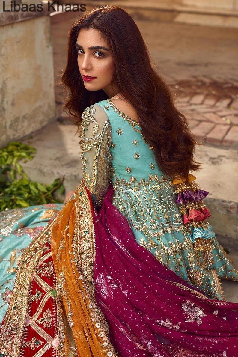 May include: A woman wearing a teal blue and gold embroidered dress with a long, flowing burgundy and orange dupatta. The dupatta has gold embroidery and tassels. The dress has a deep neckline and long sleeves. The woman is looking over her shoulder and has long, dark hair.