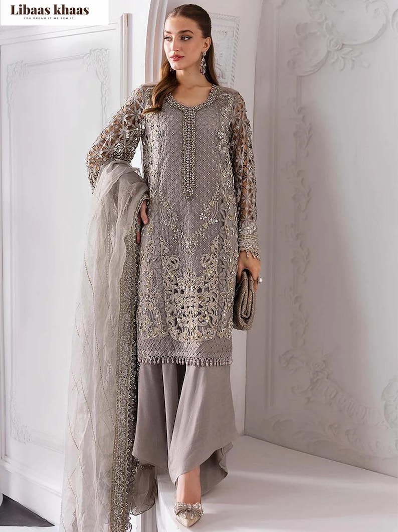 May include: A gray embroidered formal dress with long sleeves and matching pants. The dress features intricate gold detailing and a beaded neckline. A matching scarf and clutch complete the ensemble. The brand name "Libaas Khaas" is visible in the top left corner.