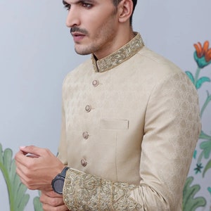 K&ouml;nnte beinhalten: Ein beigefarbener Blazer mit goldener Stickerei an Kragen und &Auml;rmeln. Der Blazer hat einen Stehkragen und einen Knopfverschluss.